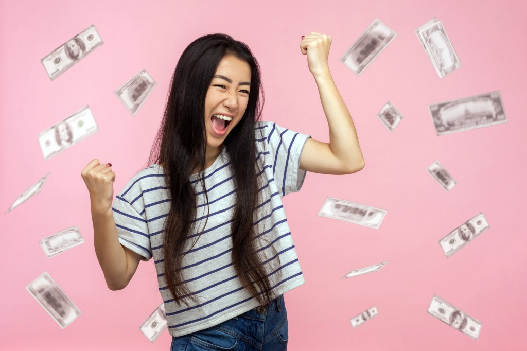 Ecstatic woman with raised punches standing in a shower falling dollars, screaming out of joy on a pink background